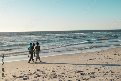 African american man piggybacking son and walking with wife at beach by beautiful sea against sky