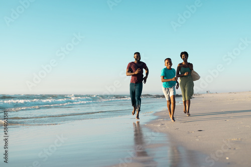 African american father and mother running behind playful son at shore against clear sky