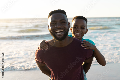 Portrait of cheerful african american man piggybacking son against scenic sea and sky at sunset