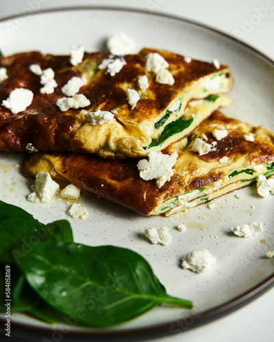 Close up of Folded omelette stuffed with spinach and feta on the white plate with spinach leaves on back. Healthy and dietary breakfast dish. Minimalistic food photo. White marble background. 