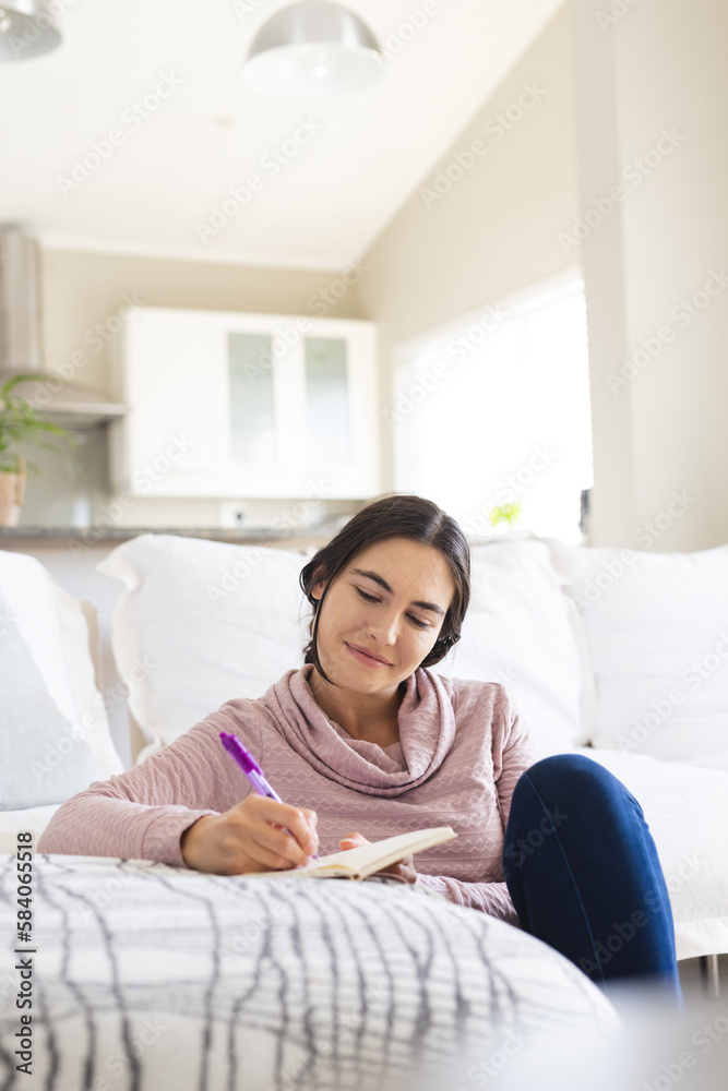 Caucasian smiling young woman thinking and writing in notepad while relaxing by sofa in living room Caucasian smiling young woman thinking and writing in notepad while relaxing by sofa in living room