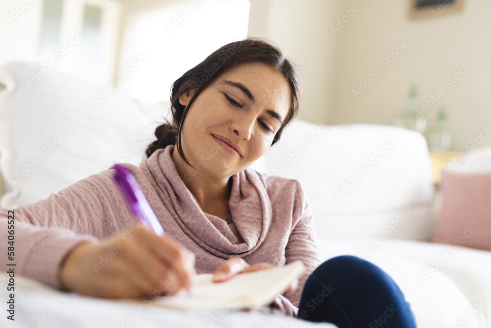 Beautiful caucasian woman thinking and writing in notepad while relaxing by sofa in living room Beautiful caucasian woman thinking and writing in notepad while relaxing by sofa in living room