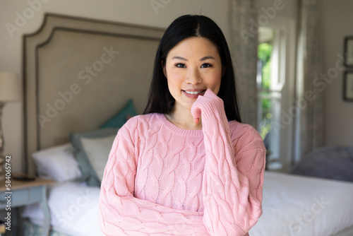 Portrait of happy asian woman wearing pink sweater smiling to camera in bedroom at home