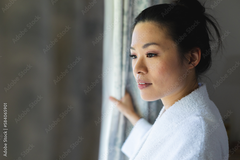 Happy asian woman wearing bathrobe, opening curtain and looking through window in bedroom Happy asian woman wearing bathrobe, opening curtain and looking through window in bedroom