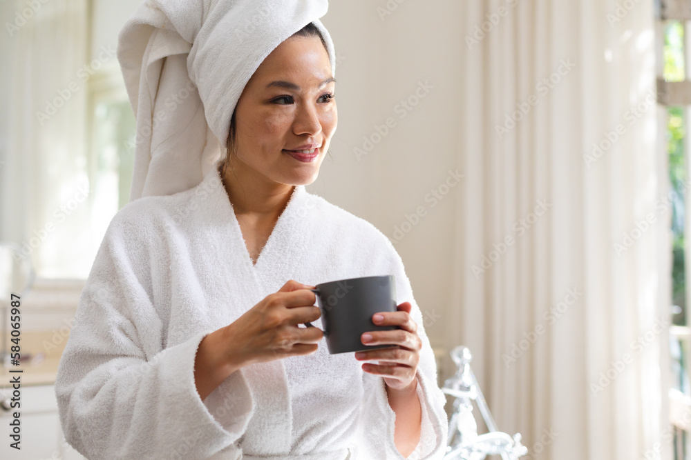 Happy asian woman wearing bathrobe and towel on head drinking coffee in bathroom Happy asian woman wearing bathrobe and towel on head drinking coffee in bathroom