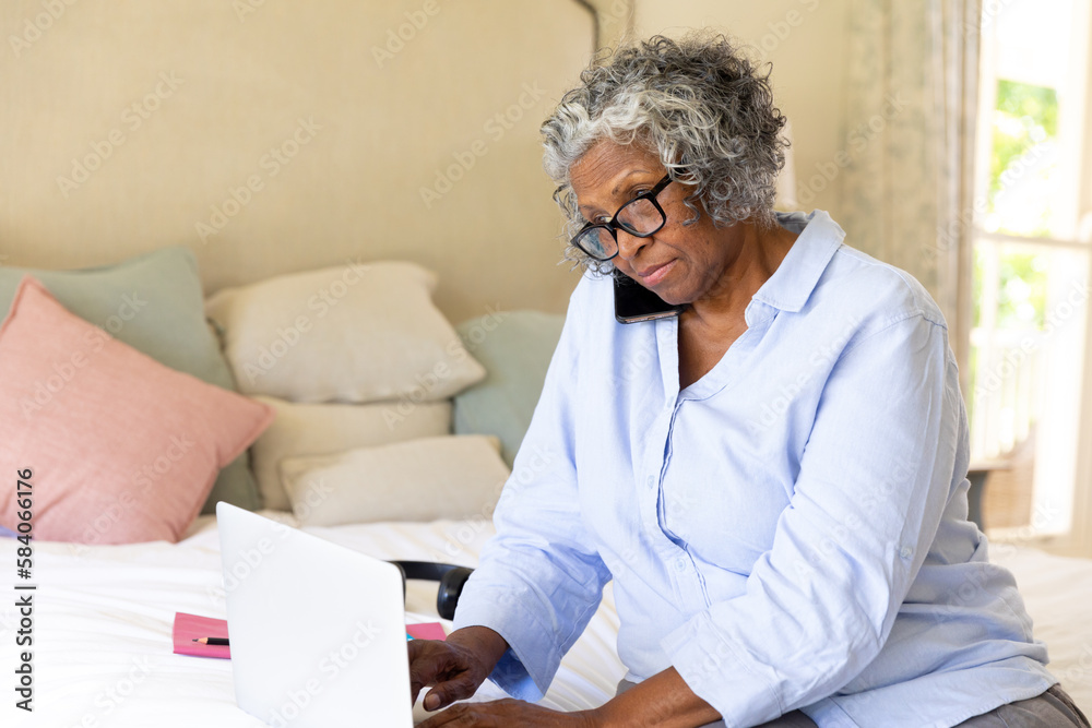 African american senior focused businesswoman talking over smartphone and using laptop on bed African american senior focused businesswoman talking over smartphone and using laptop on bed