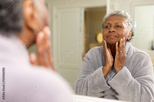 African american senior woman with short gray hair touching her face and looking in mirror at home