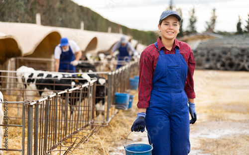 Young female farmer in overalls carrying bucket of water to get calves drinking during work on farm