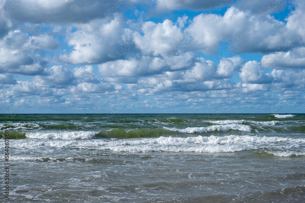 The play of white waves of the sea with clouds in the blue sky.  The rolling sea on a cloudy day.