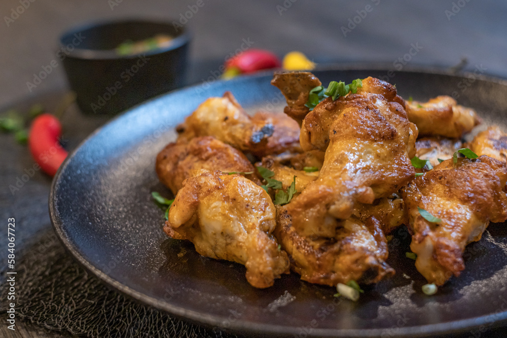Pieces of grilled chicken with greens on a black plate. Sauce in a black dish and colorful chili peppers in the background Pieces of grilled chicken with greens on a black plate. Sauce in a black dish and colorful chili peppers in the background