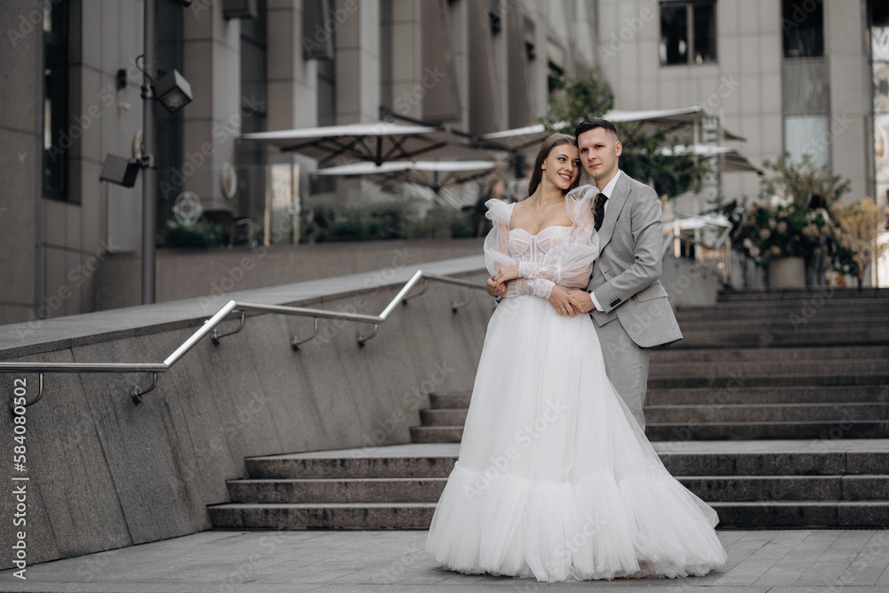 Full length portrait of just married couple, posing outdoors. Bride standing with closed eyes, smiling, groom huggs bride. Concept of love and marriage. Full length portrait of just married couple, posing outdoors. Bride standing with closed eyes, smiling, groom huggs bride. Concept of love and marriage.