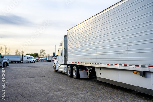 Big rig white semi truck with refrigerated semi trailer driving on the truck stop parking lot along another semi trucks looking for free parking space photo