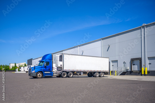 Bright blue day cab big rig semi truck with roof spoiler and refrigerator semi trailer standing in warehouse loading dock waiting for the next load