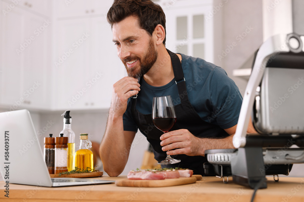 Man with glass of wine making dinner while watching online cooking course via laptop in kitchen Man with glass of wine making dinner while watching online cooking course via laptop in kitchen