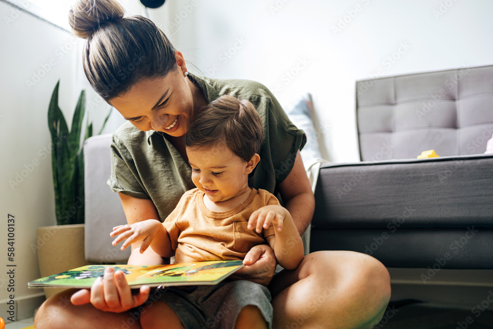 Mom reading a book with baby boy at home. Early age children education, development. Mother and child spending time together. Candid lifestyle. - obrazy, fototapety, plakaty 