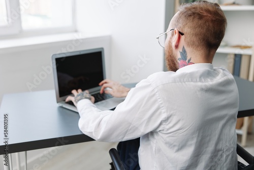 A man wheelchair businessman with tattoos in the office works at a laptop online, business process, a wheelchair close-up, integration into society, the concept of working a person with disabilities