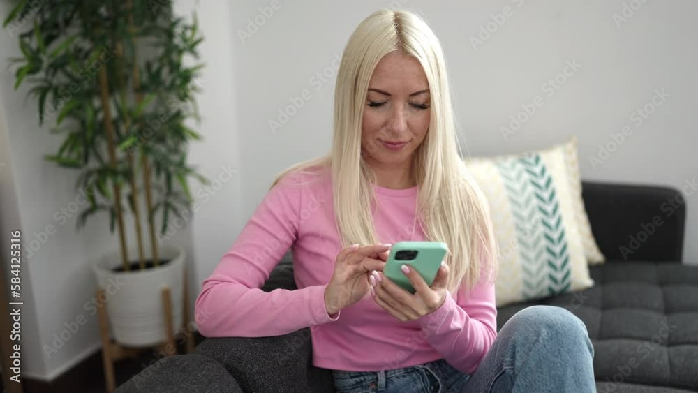 Young blonde woman using smartphone doing ok gesture at home Young blonde woman using smartphone doing ok gesture at home