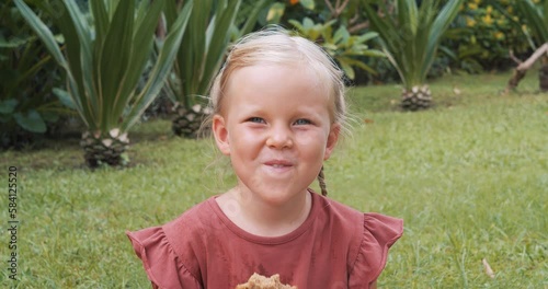 Little smiling girl eating a slice of banana muffin and looking at the camera outdoors.