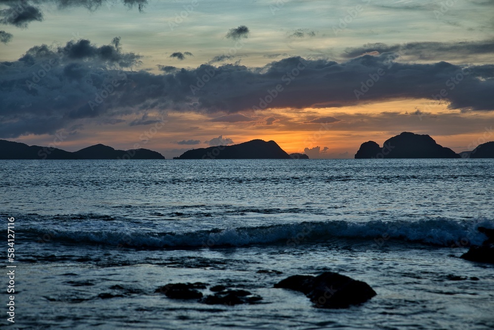 Picturesque sunset at the sea of El Nido, Palawan in the Philippines, the whole sky glows in golden orange and yellow tones, hills in the background.