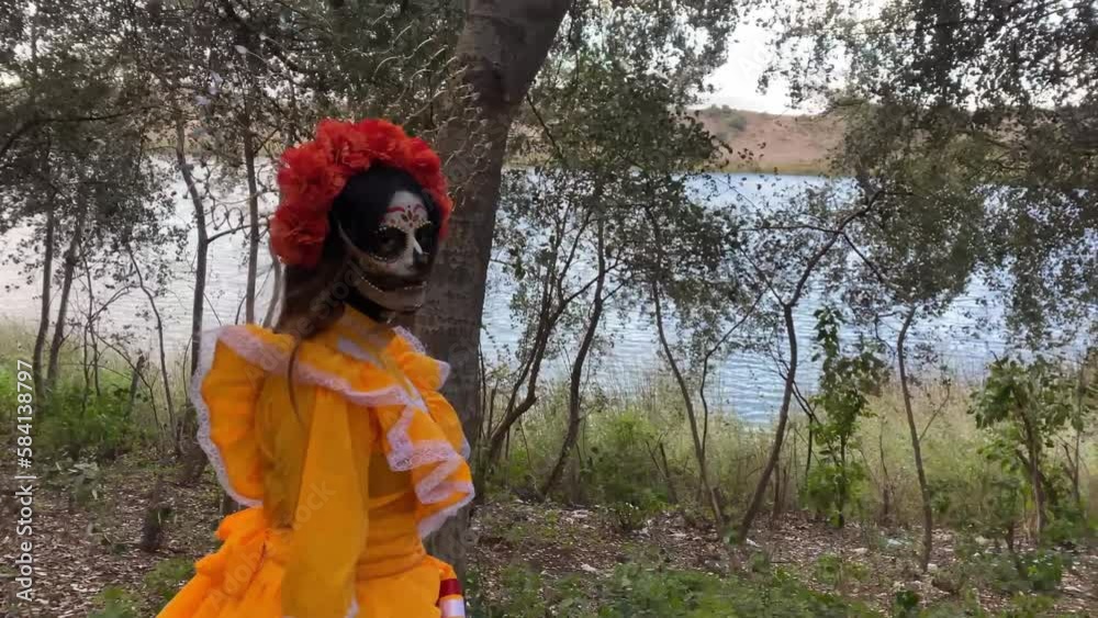 Woman in a Mexican Day of the Dead orange costume moving through a forest with a lake behind her Woman in a Mexican Day of the Dead orange costume moving through a forest with a lake behind her