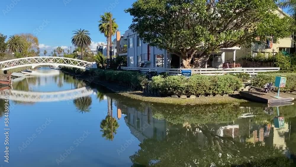 Slow left to right pan of the peaceful and serene waterways of the Venice Canals. The trees and houses line the waterway and their reflection is clearly seen in the water on a sunny clear day. Slow left to right pan of the peaceful and serene waterways of the Venice Canals. The trees and houses line the waterway and their reflection is clearly seen in the water on a sunny clear day.