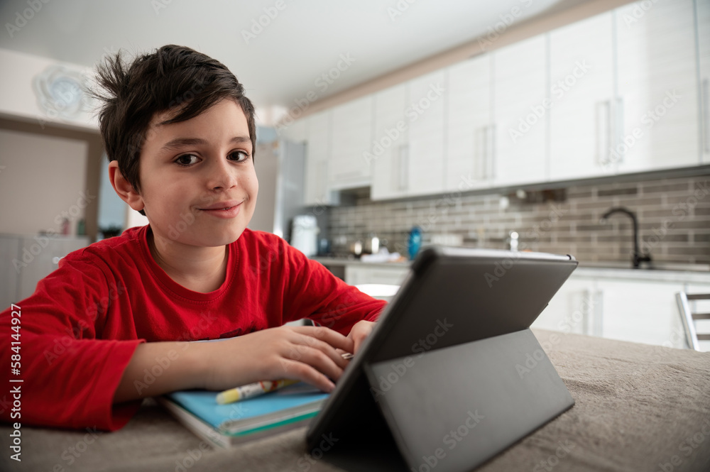 Elementary school student boy on distance learning, smiling looking at camera, doing homework, studying online, watching webinar on digital tablet, holding a pen and making notes in his textbook Elementary school student boy on distance learning, smiling looking at camera, doing homework, studying online, watching webinar on digital tablet, holding a pen and making notes in his textbook