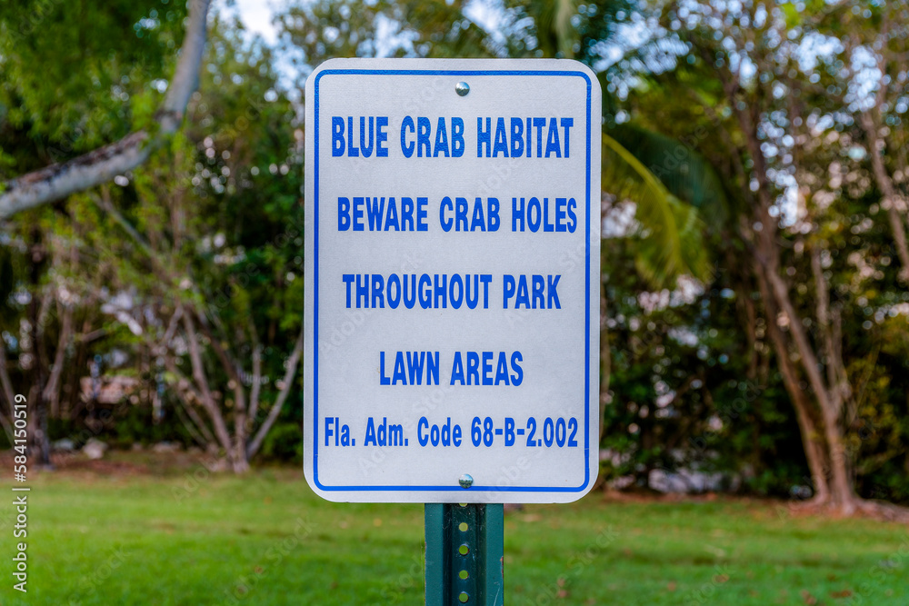 Sign post with Blue Crab Habitat Blue Crab Holes Throughout Park Lawn Areas in Miami, Florida. Close-up of a sign post against the lawn and trees in the blurred background. Sign post with Blue Crab Habitat Blue Crab Holes Throughout Park Lawn Areas in Miami, Florida. Close-up of a sign post against the lawn and trees in the blurred background.