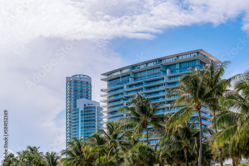 Modern high-rise glass buildings in Miami, Florida against the flat clouds in the sky. There are towers on the left near the wider building on the right behind the coconut trees at the front.