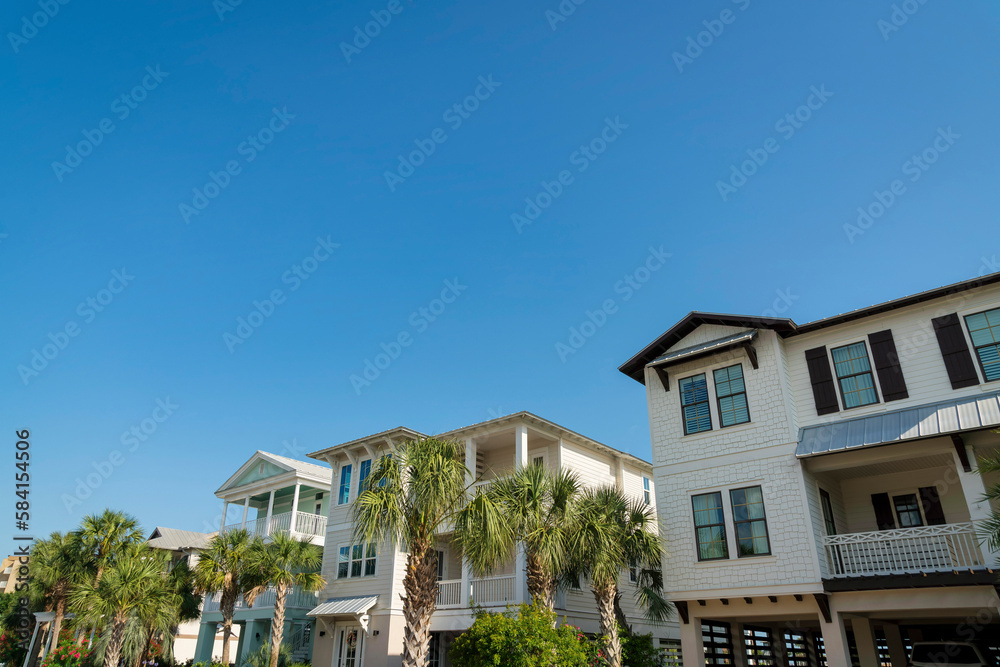 Row of three-storey houses in Destin, Florida with wood sidings and parking space under. There is a house from the right with white and brown exterior along with houses with plants and trees at front.