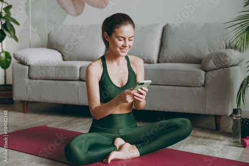 Full body smiling young strong sporty athletic fitness trainer instructor woman wear green tracksuit use mobile cell phone training do exercises at home gym indoor. Workout sport motivation concept.