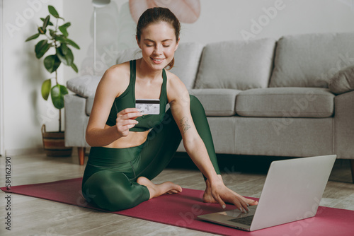 Full body fun young strong athletic fitness trainer instructor woman wear green tracksuit use laptop pc computer credit card training do exercises at home gym indoor Workout sport motivation concept