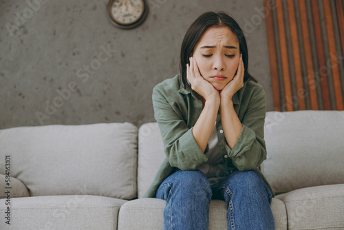 Depressed young upset sad woman of Asian ethnicity wearing casual clothes holding prop up face sits on grey sofa couch stay at home hotel flat rest relax spend free spare time in living room indoor.