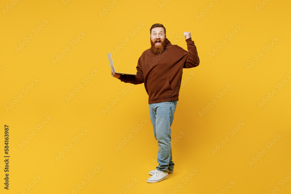 Full body side view surprised young redhead IT man wear brown hoody casual clothes hold use work on laptop pc computer do winner gesture isolated on plain yellow background studio Lifestyle concept Full body side view surprised young redhead IT man wear brown hoody casual clothes hold use work on laptop pc computer do winner gesture isolated on plain yellow background studio Lifestyle concept