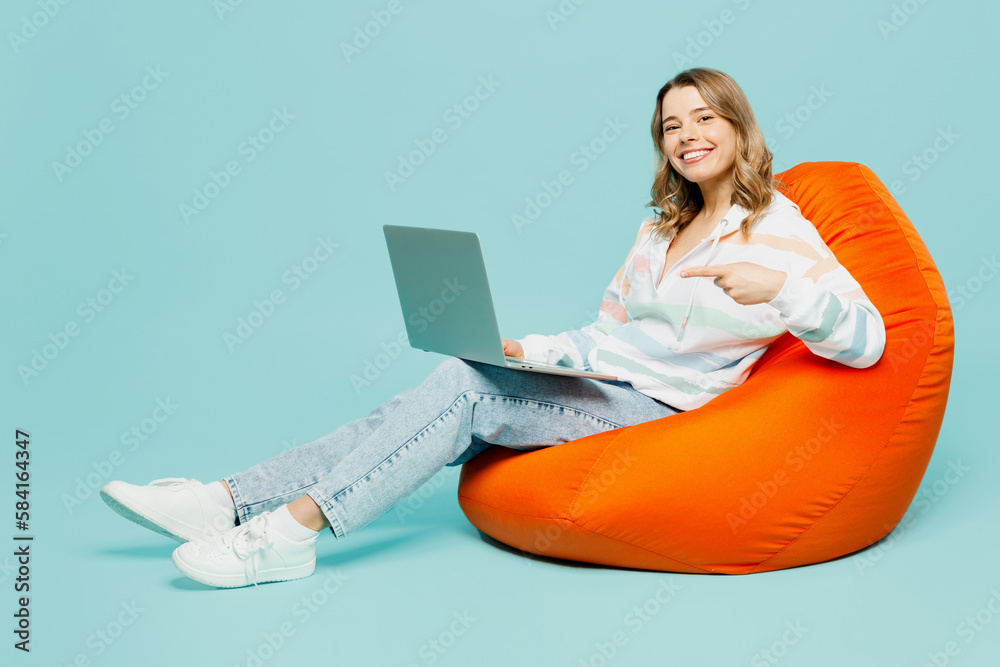 Naklejka premium Full body young IT smiling fun woman wear striped hoody sit in bag chair hold use work point finger on laptop pc computer isolated on plain pastel light blue cyan background studio Lifestyle concept