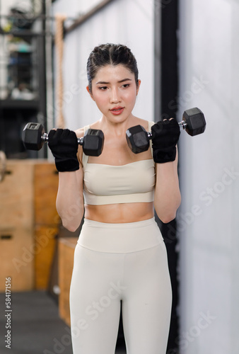 Asian young female fit strong body sporty athletic fitness model in sport bra legging gloves standing posing holding lifting metal dumbbell weight training arms exercise at CrossFit gym.