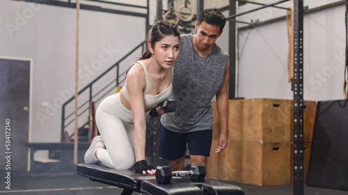 Asian professional muscular male personal trainer teaching female fit strong body sporty athletic fitness model in sport bra legging and gloves lifting dumbbells exercising triceps in CrossFit gym