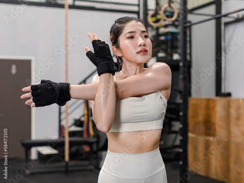 Asian young muscular fit strong body sporty athletic sexy female fitness workout sports bra leggings gloves concentrate standing holding hands arms stretching in Crossfit gym with equipment