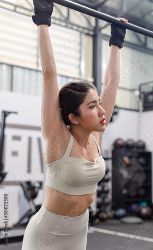 Asian young female fit strong body sporty athletic fitness model in sport bra legging and gloves hanging sweating stretching on metal gymnastic bar take break breathing after exercise training in gym