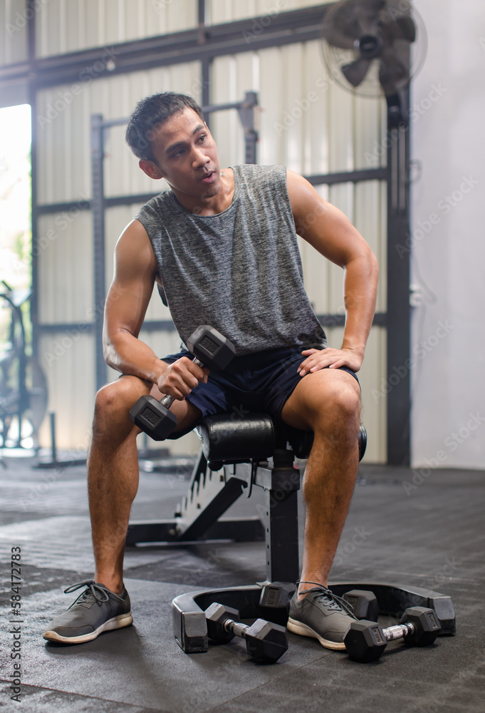 Obraz premium Asian young muscular fit strong body sporty athletic male fitness model in workout sleeveless sportswear holding lifting metal dumbbell weight training arms exercise at CrossFit gym