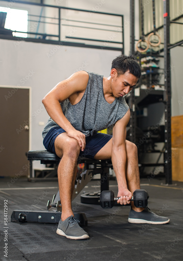 Asian young muscular fit strong body sporty athletic male fitness model in workout sleeveless sportswear holding lifting metal dumbbell weight training arms exercise at CrossFit gym Asian young muscular fit strong body sporty athletic male fitness model in workout sleeveless sportswear holding lifting metal dumbbell weight training arms exercise at CrossFit gym