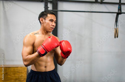 Asian young muscular fit strong body sporty athletic shirtless male fitness model in red boxing gloves with tattoo standing guarding footwork workout exercising sparring training alone in Crossfit gym
