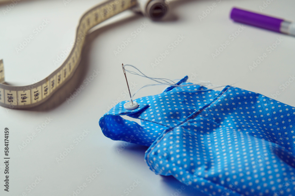 Small cute started blue polka dot costume with needle overalls for toy near measure tape and pen on white table. Sewing process of stuffed toys and clothes. Knitting hobby. DIY, children store Small cute started blue polka dot costume with needle overalls for toy near measure tape and pen on white table. Sewing process of stuffed toys and clothes. Knitting hobby. DIY, children store