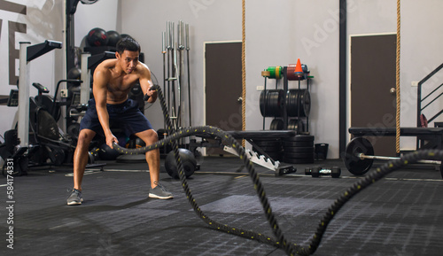 Asian young muscular fit strong body sporty athletic sexy shirtless male fitness with six pack abs standing holding battle ropes working out exercising training alone in Crossfitgym full of equipment photo