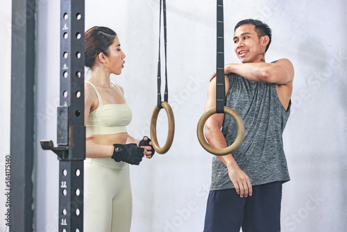 Asian young professional muscular male personal trainer teaching female fit strong body sporty athletic fitness model in sport bra legging and gloves using gymnastic hanging rings in crossfit gym