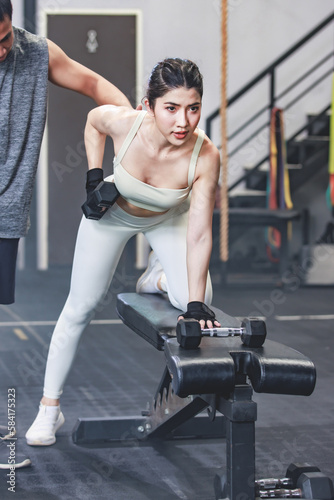 Asian professional muscular male personal trainer teaching female fit strong body sporty athletic fitness model in sport bra legging and gloves lifting dumbbells exercising triceps in CrossFit gym