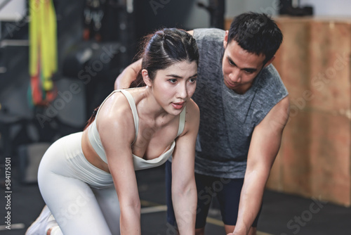 Asian professional muscular male personal trainer teaching female fit strong body sporty athletic fitness model in sport bra legging and gloves lifting dumbbells exercising triceps in CrossFit gym