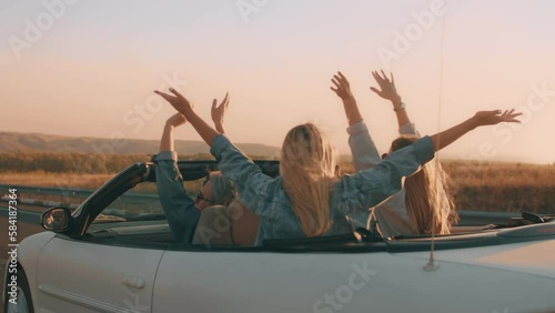 Young people in a white car with open top drive at sunset along a country road and raise their hands in the air in pleasure. Group of young people enjoys a vacation in a car country trip
