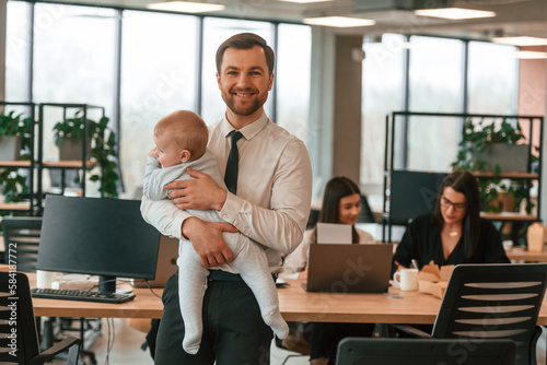 Father is smiling, holding his son. Infant baby is in the office where group of people are working together