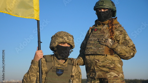 Military couple of ukrainian army with flag of Ukraine posing on camera against blue sky. Portrait of young soldiers in camouflage uniform with blue-yellow banner at countryside. Invasion resistance