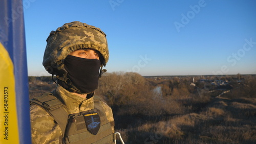 Dolly shot of male soldier in military uniform and helmet holds a waving flag of Ukraine to peak of hill. Ukrainian army man stands with lifted national banner against blue sky. Invasion resistance